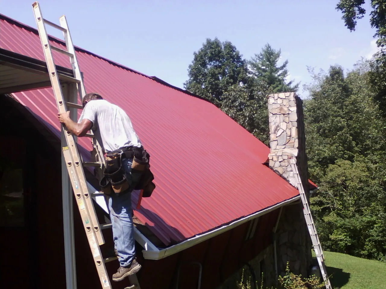 Roofer on a ladder beside a red metal roof and stone chimney — Soffit & Fascia Repair by Roofing Brothers in Mount Juliet
